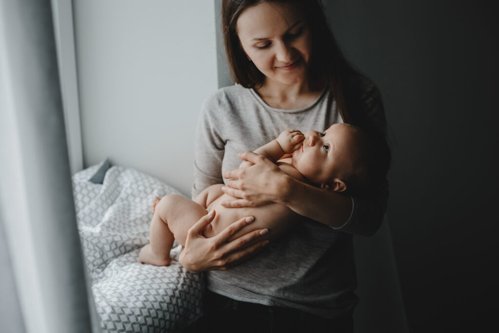 Lovely mom holds happy newborn boy standing before a bright window